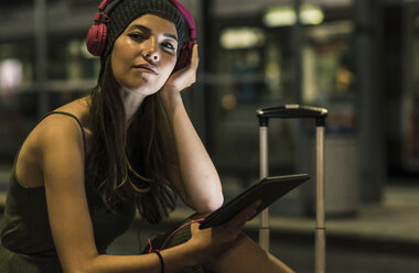 Portrait of young woman with headphones and tablet waiting at station by night - UUF11053