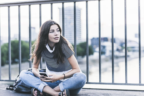 Porträt einer jungen Frau mit Mobiltelefon auf einer Brücke sitzend - UUF11059