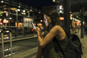 Junge Frau mit Rucksack und Gepäck, die nachts am Bahnhof wartet und ein Mobiltelefon benutzt - UUF11071