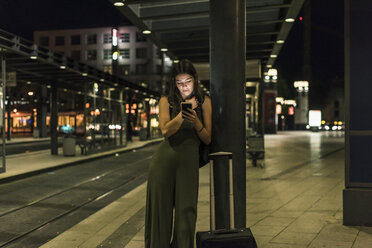 Junge Frau, die nachts am Bahnhof wartet und ein Mobiltelefon benutzt - UUF11072
