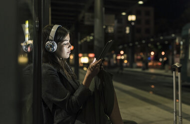 Young woman with headphones waiting at the station by night using tablet - UUF11076