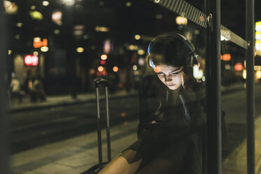 Young woman with headphones waiting at the station by night using tablet - UUF11077