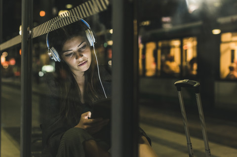 Porträt einer jungen Frau mit Kopfhörern, die nachts auf dem Bahnhof wartet und ein Tablet benutzt, lizenzfreies Stockfoto