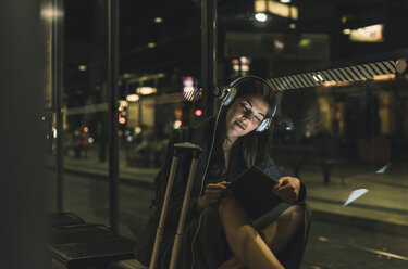 Portrait of young woman with headphones and tablet waiting at station by night - UUF11079