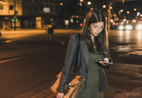 Junge Geschäftsfrau mit Ledertasche, die bei Nacht auf ihr Handy schaut - UUF11081