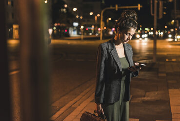 Junge Geschäftsfrau mit Ledertasche, die bei Nacht auf ihr Handy schaut - UUF11083
