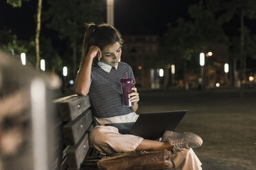 Junge Frau mit Smoothie sitzt nachts auf einer Bank und benutzt einen Laptop - UUF11086