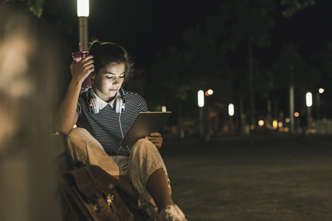 Junge Frau mit Smoothie sitzt nachts auf einer Bank und benutzt ein Tablet, lizenzfreies Stockfoto