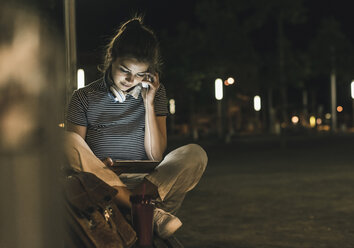 Junge Frau mit Smoothie sitzt nachts auf einer Bank und benutzt Tablet und Kopfhörer - UUF11089