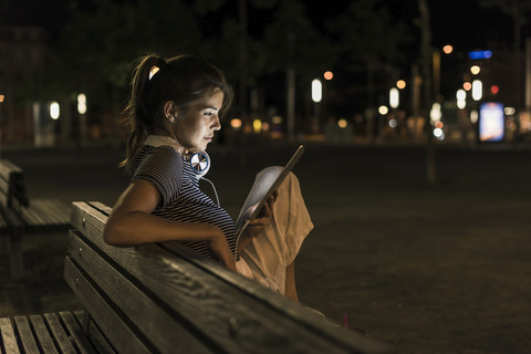 Junge Frau sitzt nachts auf einer Bank und benutzt ein Tablet, lizenzfreies Stockfoto