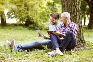 Älterer Vater sitzt neben seinem erwachsenen Sohn in einem Park mit Tablet - HAPF01865