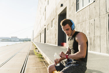 Junger Sportler mit Kopfhörern, der an einer Wand sitzt und seine Smartwatch überprüft - UUF11148