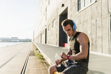 Young athlete wearing headphones, sitting on a wall, checking smartwatch - UUF11148