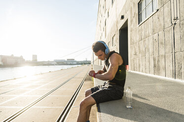 Young athlete wearing headphones, sitting on a wall, checking smartwatch - UUF11151