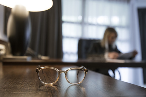 Brille auf dem Schreibtisch mit einer Geschäftsfrau, die im Hintergrund Dokumente überprüft, lizenzfreies Stockfoto