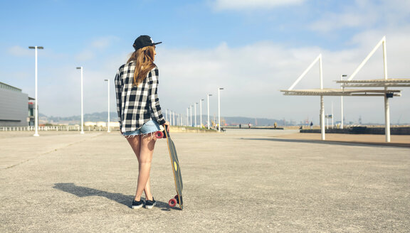 Rückenansicht einer jungen Frau mit Longboard vor einer Strandpromenade, Teilansicht - DAPF00767