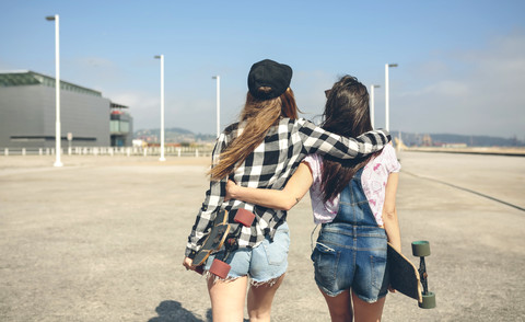 Rückenansicht von zwei jungen Frauen mit Longboards, die Arm in Arm auf der Strandpromenade gehen, lizenzfreies Stockfoto