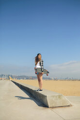 Junge Frau mit Longboard auf einer Mauer an der Strandpromenade stehend - DAPF00780