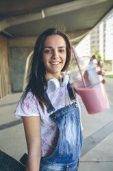 Portrait of smiling young woman with headphones showing strawberry smoothie - DAPF00792