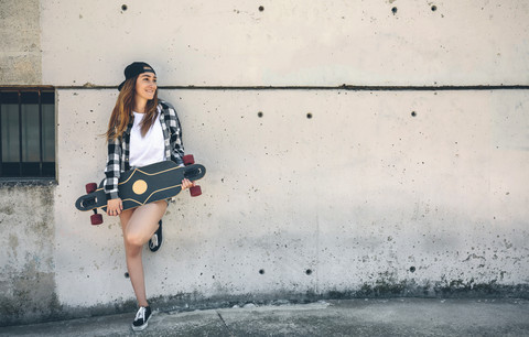 Porträt einer glücklichen jungen Frau mit Longboard vor einer Betonwand stehend, lizenzfreies Stockfoto