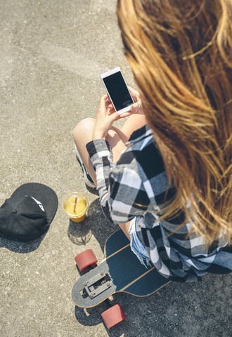 Rückenansicht einer jungen Frau mit langen Haaren, die auf einem Longboard sitzt und auf ihr Smartphone schaut, lizenzfreies Stockfoto