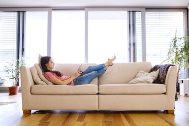 Junge Frau liegt auf der Couch und benutzt ein Tablet für einen Video-Chat zu Hause - HAPF01890