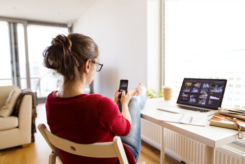 Frau sitzt zu Hause am Schreibtisch und benutzt ihr Smartphone, lizenzfreies Stockfoto