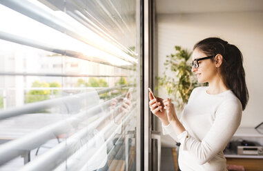 Smiling woman standing at window at home using smartphone - HAPF01936