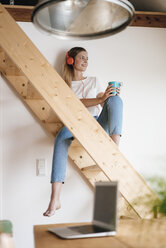 Junge Frau sitzt auf einer Holztreppe, hört Musik und trinkt Tee - GUSF00156