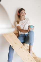 Junge Frau sitzt auf einer Holztreppe, hört Musik und trinkt Tee - GUSF00157