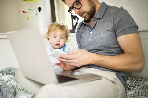 Father at home with his little son using laptop and cell phone stock photo