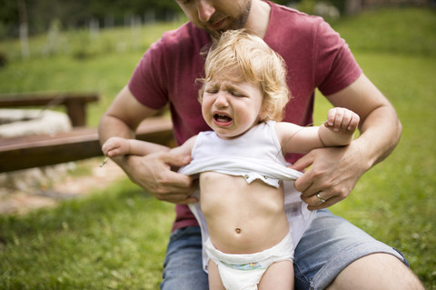 Vater, der seinen weinenden kleinen Sohn im Garten anzieht, lizenzfreies Stockfoto