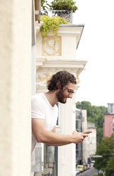 Lächelnder Mann auf dem Balkon schaut auf sein Handy - MFRF00876