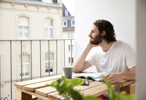 Tagträumender junger Mann sitzt mit Kaffeetasse und Zeitschrift auf dem Balkon und schaut in die Ferne - MFRF00880