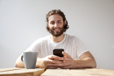 Portrait of smiling man sitting with coffee mug on balcony using cell phone - MFRF00881