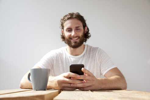 Porträt eines lächelnden Mannes, der mit einem Kaffeebecher auf einem Balkon sitzt und ein Handy benutzt - MFRF00881