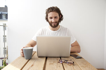 Porträt eines lächelnden Mannes, der mit einem Kaffeebecher auf einem Balkon sitzt und einen Laptop benutzt - MFRF00884
