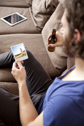 Mann sitzt mit Bierflasche auf der Couch und betrachtet ein Sofortfoto - MFRF00901