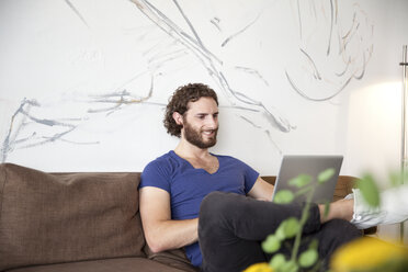 Lächelnder junger Mann sitzt auf einer Couch in einem Café und benutzt einen Laptop - MFRF00903