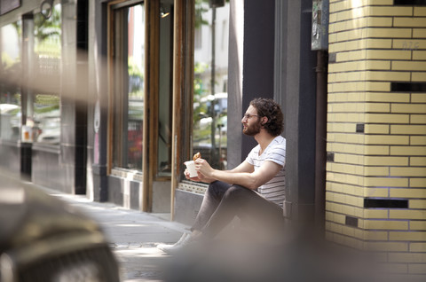 Junger Mann mit Kaffeetasse und Croissant sitzt am Eingang eines Cafés, lizenzfreies Stockfoto