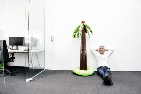 Lächelnder reifer Geschäftsmann sitzt neben einer aufblasbaren Palme im Büro - KNSF02215