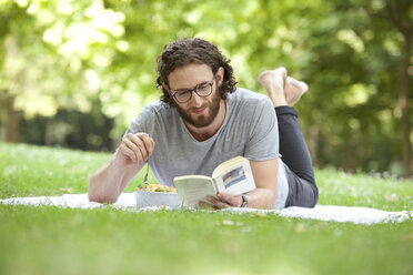 Mann liest auf einer Decke in einem Park ein Buch und isst Nudelsalat - MFRF00917