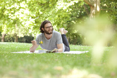 Smiling man with book eating noodle salad on blanket in a park - MFRF00918