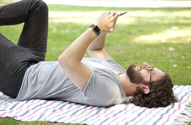 Mann liegt auf einer Decke in einem Park und benutzt ein Mobiltelefon - MFRF00923