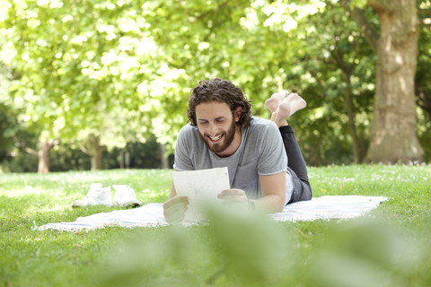 Lachender Mann, der auf einer Decke in einem Park liegt und einen Brief liest, lizenzfreies Stockfoto