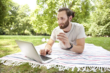 Mann mit Kaffee zum Mitnehmen, auf einer Decke im Park liegend, mit Laptop - MFRF00930