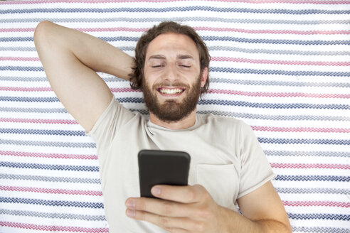 Porträt eines lächelnden Mannes, der in einem Park auf einer Decke liegt und ein Handy benutzt, Ansicht von oben - MFRF00932