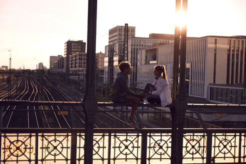 Deutschland, München, Junges Paar sitzt auf Brücke und genießt Sonnenuntergang - SUF00263