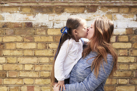 Mother kissing her little daughter in front of brick wall stock photo