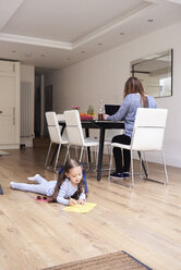 Kleines Mädchen liegt auf dem Boden und malt ein Bild, während ihre Mutter im Hintergrund am Laptop arbeitet - IGGF00014
